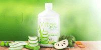 Bottle of aloe vera juice with sliced aloe and fruit on a table, green blurred background.